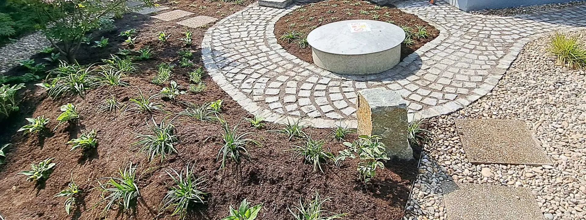 Neu angelegte Gartenfläche mit Stauden, geschwungenem Pflasterweg und rundem Betonelement.