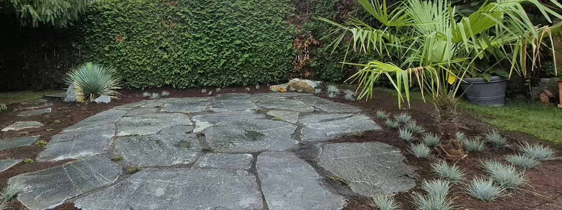 Großzügige Natursteinfläche im Garten, umrahmt von Stauden, Gräsern, Palmen und immergrüner Hecke